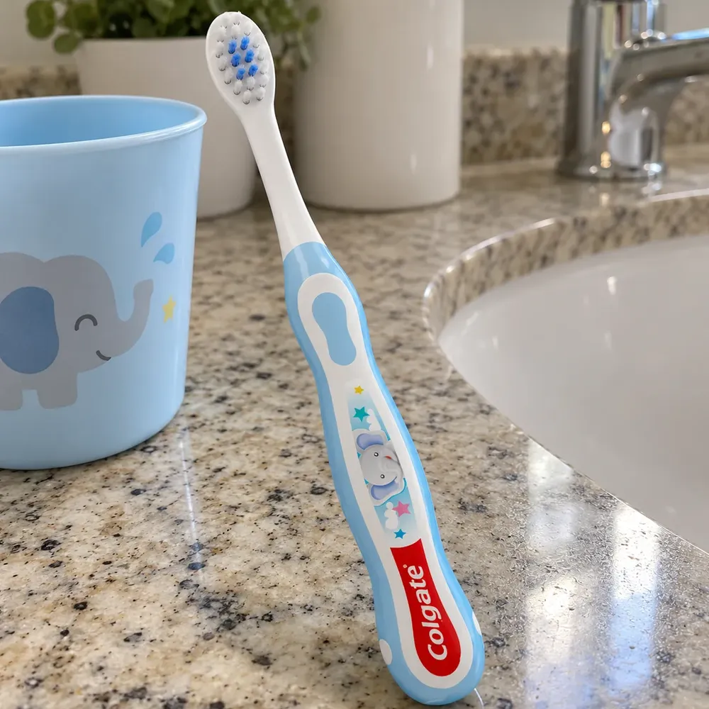 Escova Colgate My First azul com desenho de elefante, sem a cartela, em pé na bancada de granito do banheiro, ao lado de um copo infantil e da pia