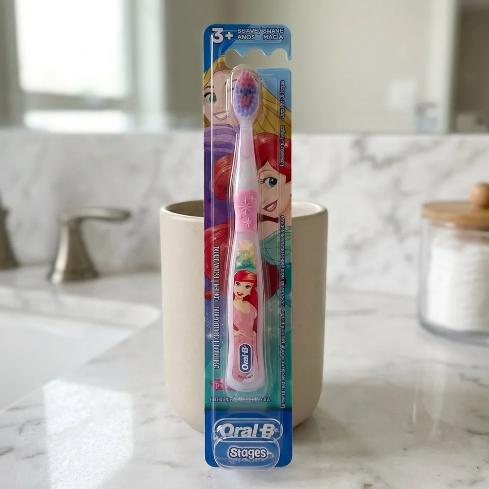 Escova Oral-B Stages rosa de princesas, na cartela marcada 3+, apoiada num copo bege sobre a pia de mármore do banheiro